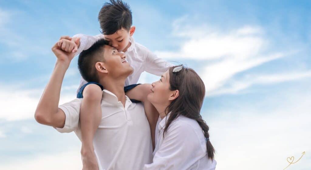 família composta pelo pai, mãe e filho. Ambos com camiseta branca. Mãe e pai abraçados olhando para o filho que está sentado nos ombros do pai.
