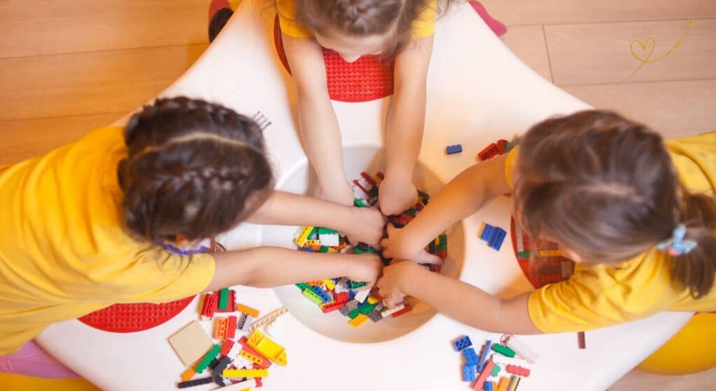 Três crianças brincando em uma mesa branca com peças de montar coloridas.