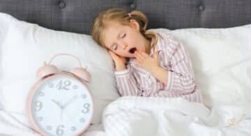 rotina-noturna Menina bocejando usando um pijama comprido com listrar rosa claro e branco, deitada em um cama com um relógio analógico grande rosa e branco.