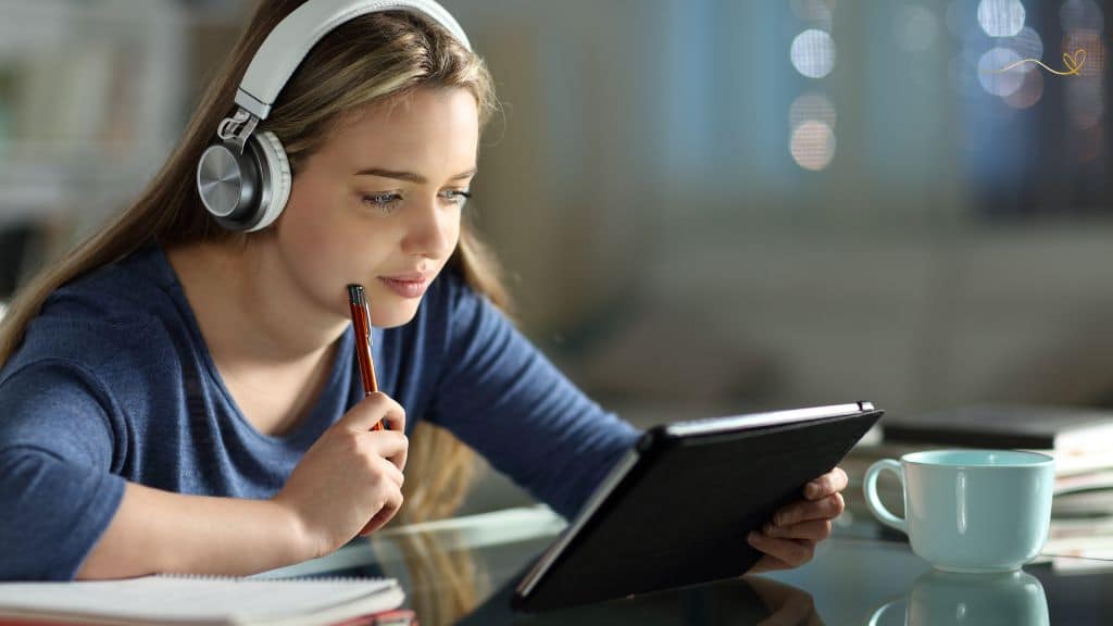 Adolescência e Tecnologia Menina de cabelos compridos loira, usando fones de ouvido brancos e segurando um tablet com a mão esquerda e com a mão direita segura uma caneta vermelha apoiada no queixo. Com os braços e mãos apoiados em uma mesa de vidro contendo uma xícara branca.