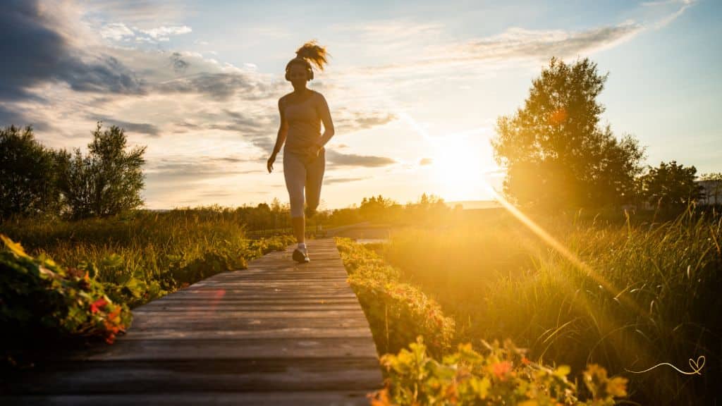 caminho em madeira, rodeado por vegetação formando um lindo corredor ao pôr-do-sol, com uma mulher vestindo roupas de corrida, cabelo preso e fone de ouvido, correndo por esse caminho.