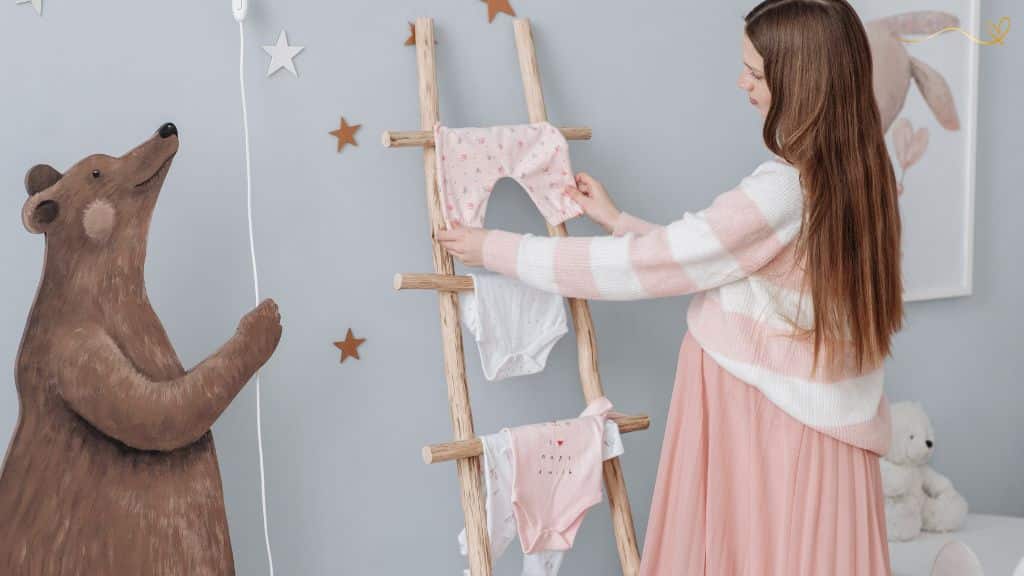 organizando o enxoval do bebê Mãe gestante organizando quarto da bebê, com um guarda-roupa decorado com um urso grande e estrelas douradas, uma escadinha de madeira com peças de roupas rosas.