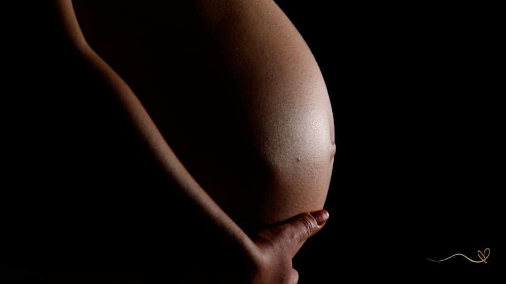 sintomas incomuns da gravidez gestante segurando a barriga com a mão direita em perfil com fundo escuro da imagem.
