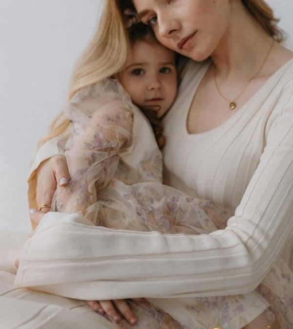 Mãe e filha usando roupas em tons pastel, abraçadas com carinho em um ambiente calmo e iluminado.