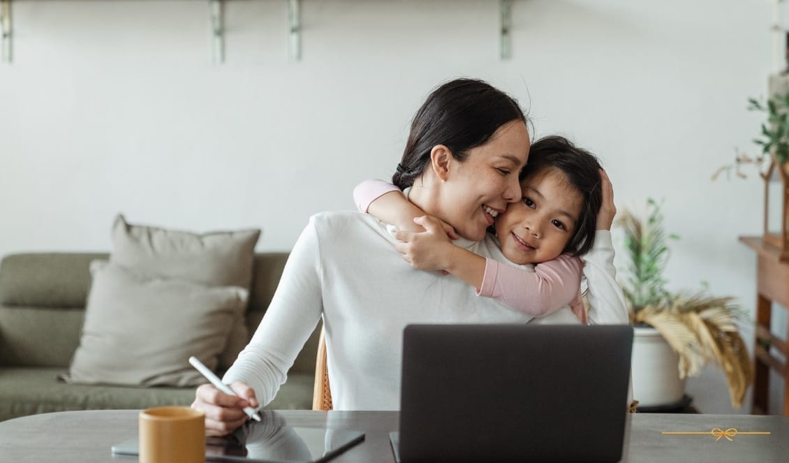 mulher abraçando sua filha, sentada a mesa com seu notebook e tablet, durante trabalho em casa.